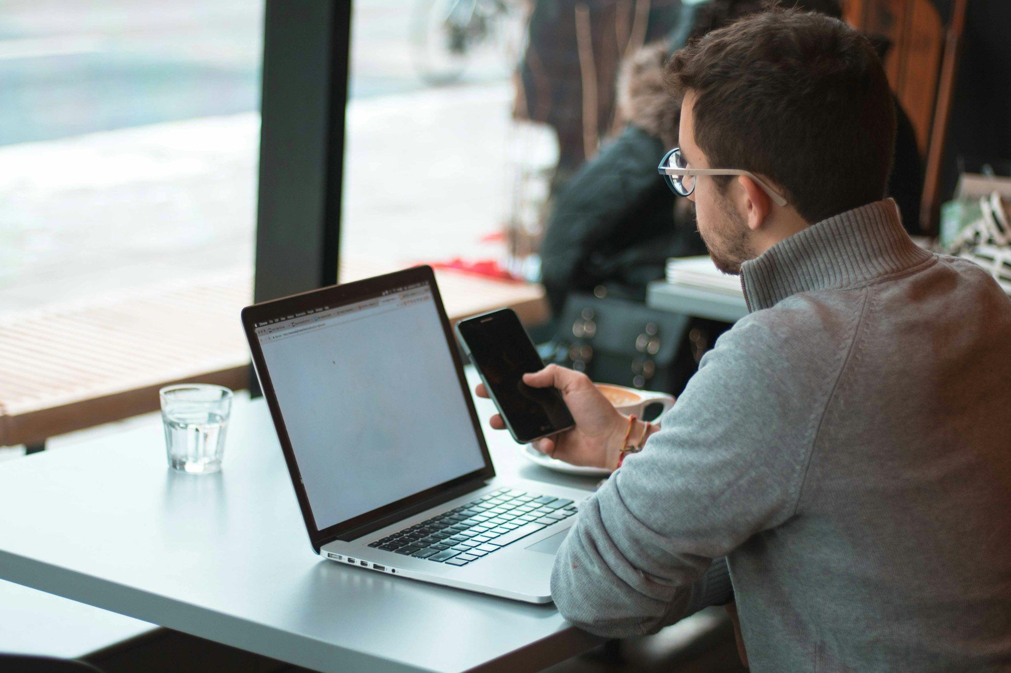 A man at a table by a window with a laptop, looking at his phone