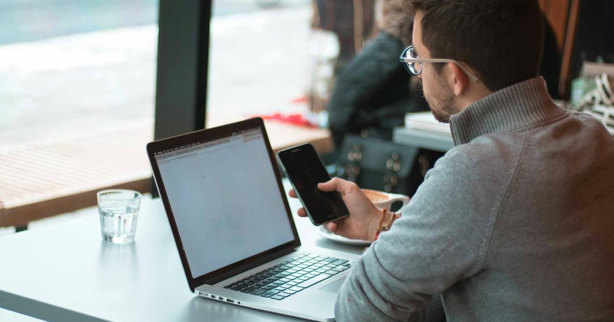 A man at a table by a window with a laptop, looking at his phone